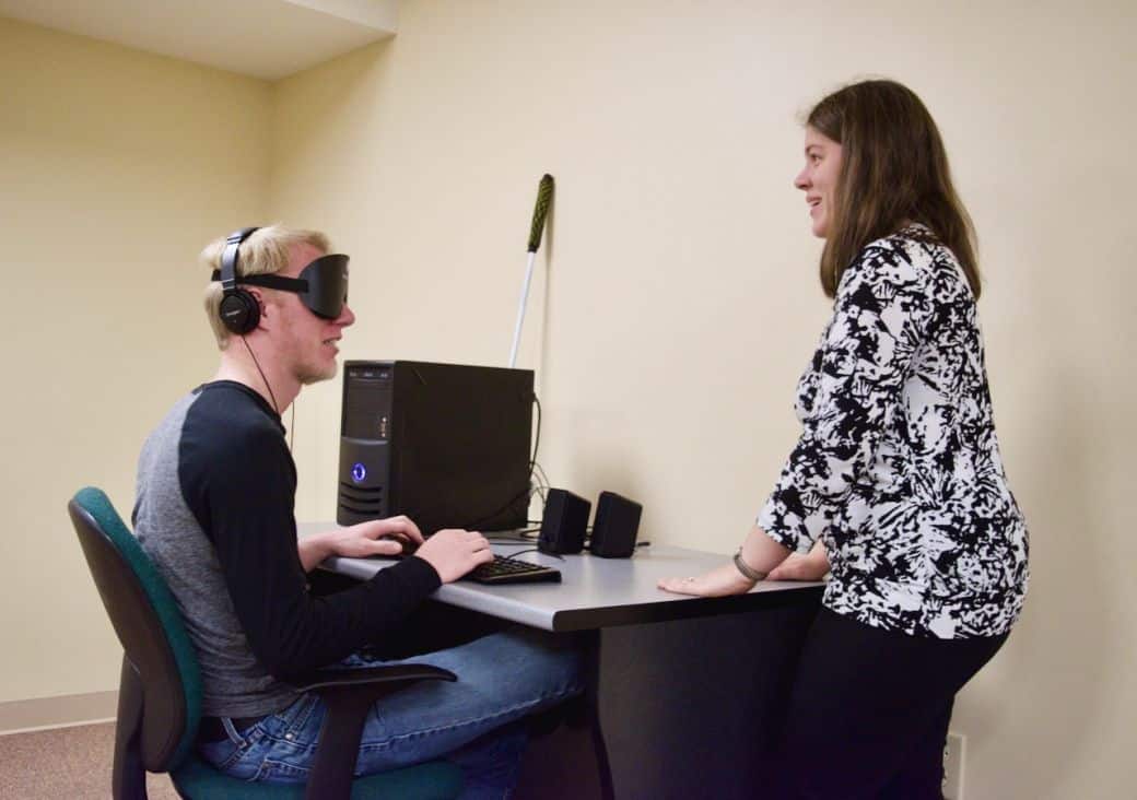 Student at the Colorado Center for the Blind (CCB) sits at a computer with no monitor, wearing thick black masks and headphones